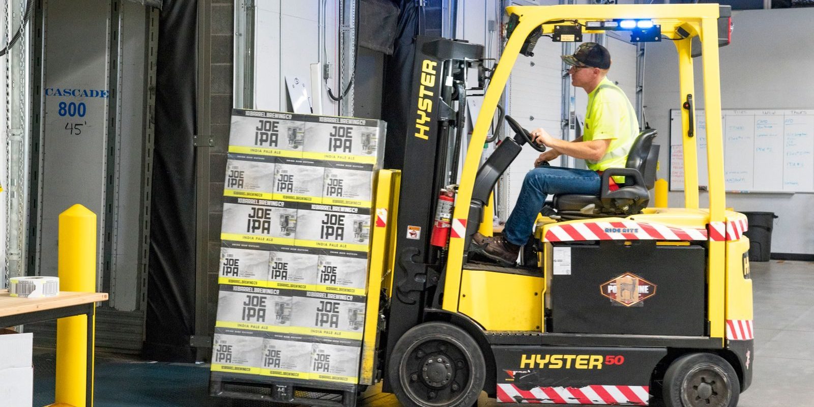A worker drives a Hyster forklift moving Joe IPA boxes in a warehouse.
