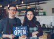 Friendly café owners holding an 'Open' sign welcoming customers into their modern coffee shop.