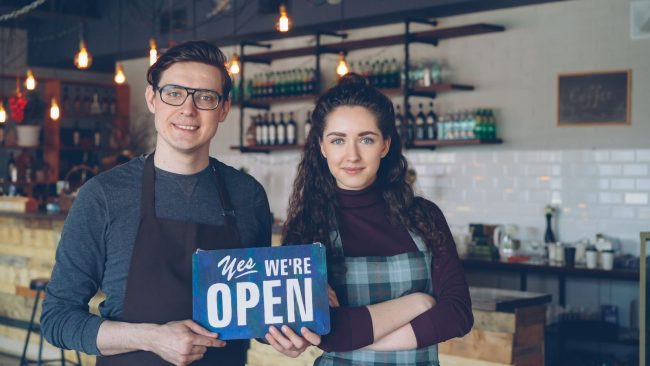 Pexels Photo by Vitaly Gariev Friendly café owners holding an 'Open' sign welcoming customers into their modern coffee shop.