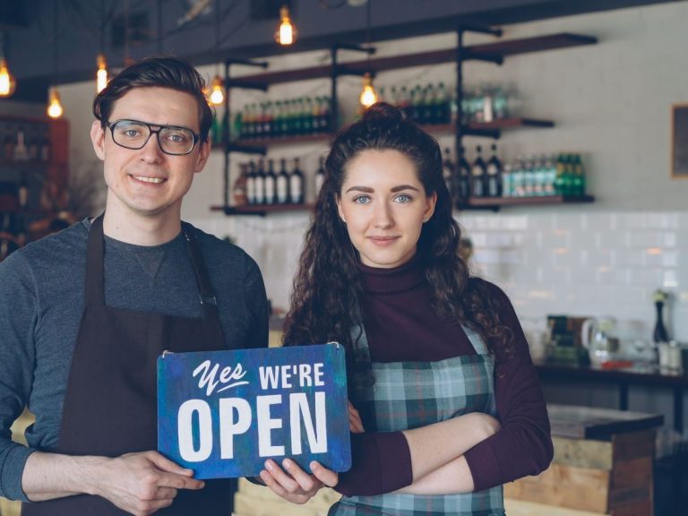 Friendly café owners holding an 'Open' sign welcoming customers into their modern coffee shop.